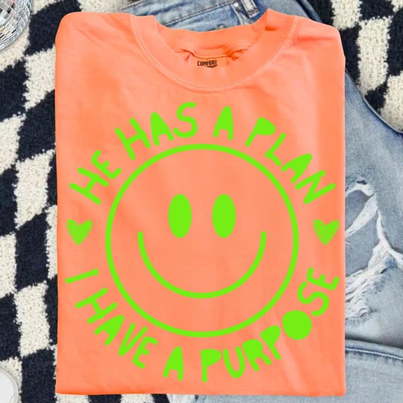 Puede incluir: Una camiseta de color coral con un dise&ntilde;o de carita sonriente verde ne&oacute;n. El texto alrededor de la carita sonriente dice "HE HAS A PLAN, I HAVE A PURPOSE". La camiseta est&aacute; doblada y colocada sobre unos vaqueros azules.
