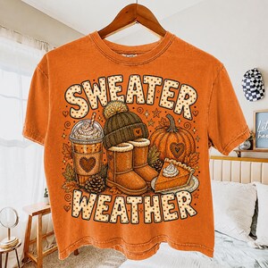 May include: An orange short-sleeve t-shirt featuring a graphic print with the text "SWEATER WEATHER" in a dotted font. The design includes a coffee cup, a knit beanie, a pair of boots, a pumpkin, and a slice of pumpkin pie with whipped cream.