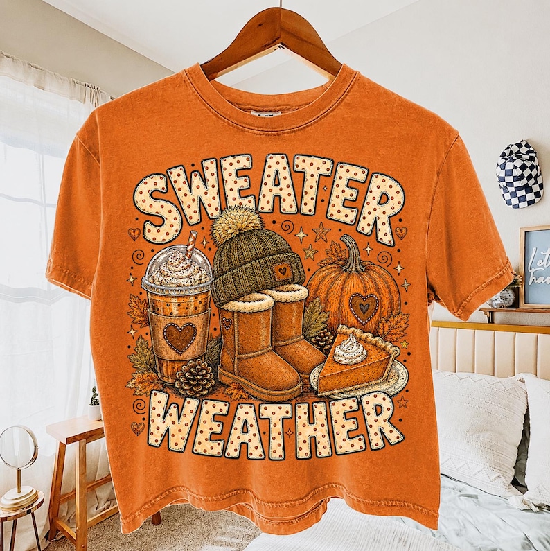 May include: An orange short-sleeve t-shirt featuring a graphic print with the text "SWEATER WEATHER" in a dotted font. The design includes a coffee cup, a knit beanie, a pair of boots, a pumpkin, and a slice of pumpkin pie with whipped cream.