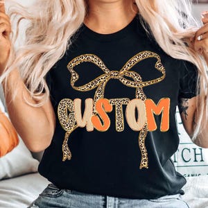 May include: Black t-shirt featuring the word "CUSTOM" in a playful design. The word "CUSTOM" is in a combination of orange and tan colors, with a leopard print bow design above it. The shirt is a casual, everyday wear item.
