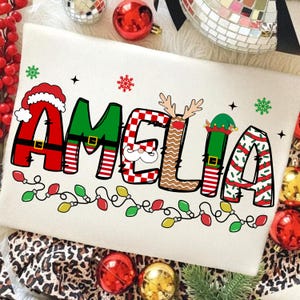 May include: A cream-colored Christmas-themed pillow with the name "AMELIA" in festive lettering. The letters are decorated with holiday elements like Santa hats, reindeer antlers, and elf hats, with a string of lights at the bottom.