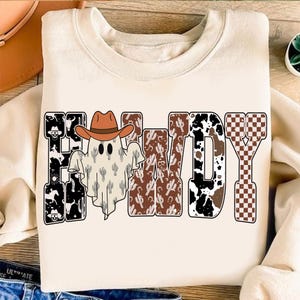 K&ouml;nnte beinhalten: Cremefarbenes Sweatshirt mit dem Wort "HOWDY" in einem Western-Design. Der Buchstabe "H" zeigt einen schwarz-wei&szlig;en Kuhdruck, und der Buchstabe "O" hat ein Gespenst mit Cowboyhut. Der Buchstabe "W" hat ein Kaktusmuster.