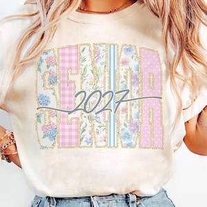 May include: Cream-colored t-shirt featuring the word "SENIOR" in a gold glitter font, with floral, gingham, and polka dot patterns. The year "2027" is written in a gray script font across the design. A stylish tee for graduation.