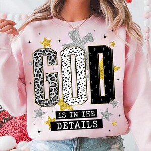Op de afbeelding: Roze sweatshirt met de tekst "GOD IS IN THE DETAILS". Het woord "GOD" is ontworpen met luipaardprint, stippen en kruisen. Gouden en zilveren sterren en een zilveren kruis sieren ook het ontwerp.