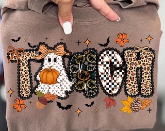 May include: Brown long-sleeve shirt featuring the word "TEACH" in a fall design. The letters are decorated with leopard print, pumpkins, a ghost holding a pumpkin, and a checkered pattern. The shirt is decorated with bats, leaves, and stars.
