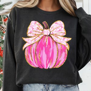 May include: Black sweatshirt featuring a pink pumpkin design with a gold glitter outline. The pumpkin has a large bow with pink ribbon accents. The design is centered on the front of the shirt.