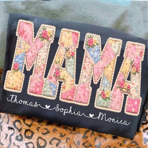 以下が含まれることがあります： パッチワーク文字で「MAMA」と書かれた濃い灰色のTシャツ。各文字は花柄と小さなバラのアクセントで飾られています。下には、「Thomas」、「Sophia」、「Monica」の名前が筆記体で書かれ、ハートで区切られています。