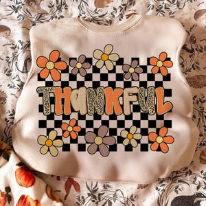 May include: Cream-colored sweatshirt with a floral and checkerboard design. The word "THANKFUL" is printed in orange with a leopard print pattern. The sweatshirt features orange, brown, and yellow flowers.