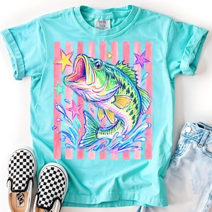 Op de afbeelding: Turquoise t-shirt met een kleurrijke illustratie van een springende vis, omringd door sterren en een roze gestreepte achtergrond. Het shirt heeft een ronde hals en opgerolde mouwen.