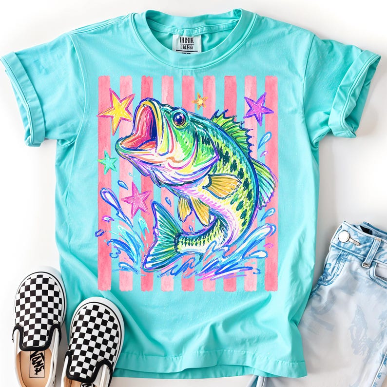 Op de afbeelding: Turquoise t-shirt met een kleurrijke illustratie van een springende vis, omringd door sterren en een roze gestreepte achtergrond. Het shirt heeft een ronde hals en opgerolde mouwen.