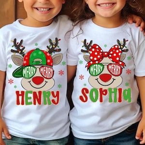 May include: White t-shirts with a Christmas reindeer design. One shirt features a green and red cap, the other a red polka dot bow. Both shirts have the text "MERRY" in sunglasses and a name below.