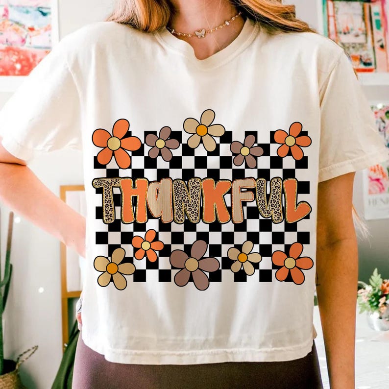 May include: Cream-colored t-shirt with the word "THANKFUL" in orange and leopard print letters. The design includes orange, brown, and yellow flowers, and a black and white checkered background.