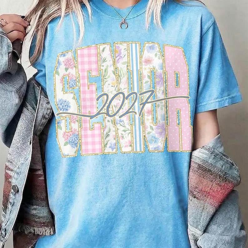 May include: Light blue t-shirt with the word "SENIOR" in a decorative font, featuring floral, plaid, and polka dot patterns. The year "2027" is printed in a gray script across the design. The shirt has short sleeves and a crew neck.