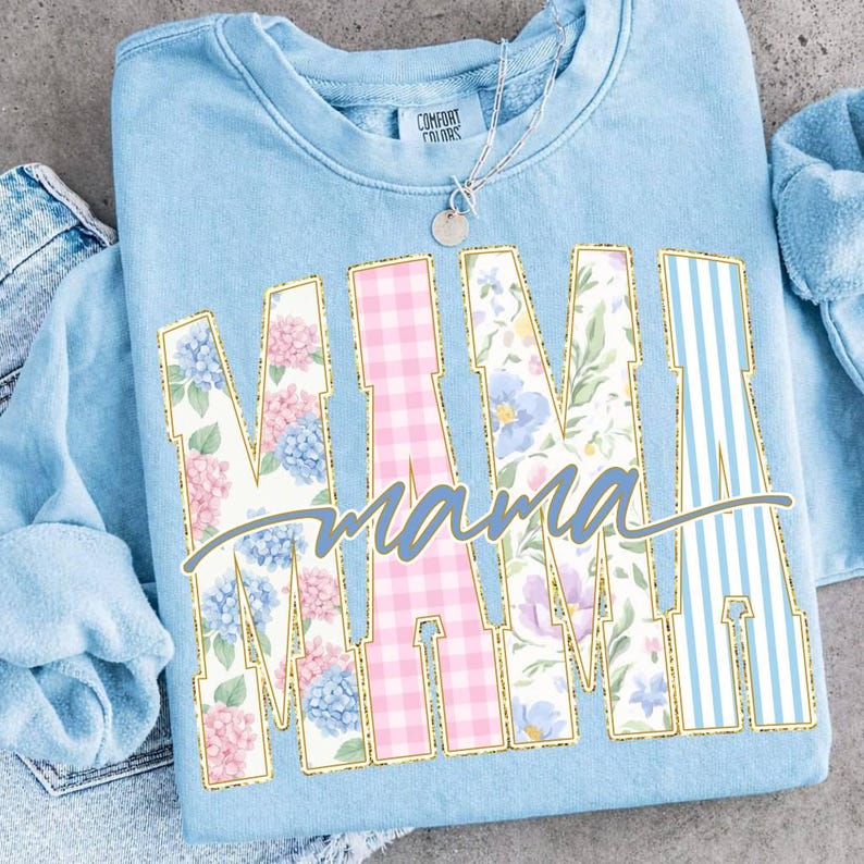 May include: Light blue sweatshirt with the word "MAMA" in large, decorative letters. The letters are filled with floral, gingham, and striped patterns, outlined in gold. The word "mama" is written in cursive below the letters. A silver necklace with a pendant is visible.