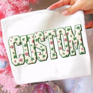 May include: White rectangular item with the word "CUSTOM" embroidered in a green and cream color scheme. The letters are filled with green and pink shamrocks. The item is held by a hand, with a pink and white background.