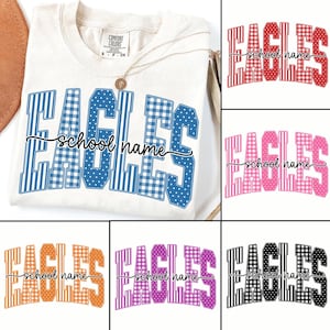 Pode incluir: Uma t-shirt cor de creme com a palavra EAGLES em letras grandes com padrões variados, incluindo xadrez vichy, riscas e pontos. O texto school name está escrito em cursivo preto sobre o centro das letras.