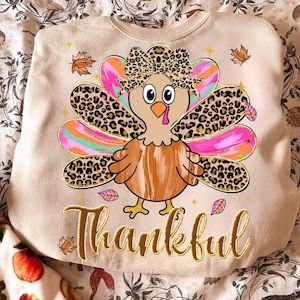 May include: A beige sweatshirt with a cartoon turkey design. The turkey has a leopard print bow and tail feathers. The word "Thankful" is written in gold script below the turkey, with autumn leaves around it.