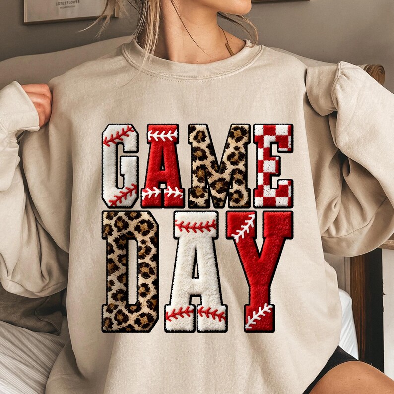 May include: A beige sweatshirt with the words "GAME DAY" in a baseball-themed design. The letters are a mix of white with red stitching, leopard print, and red and white checkered patterns, creating a sporty and eye-catching look.