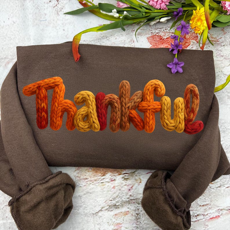 May include: A brown sweatshirt with the word "Thankful" in a knitted, textured font. The letters are in shades of orange, yellow, and red. The sleeves are folded, and the background includes flowers.