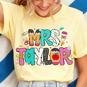 Puede incluir: Una camiseta amarilla clara con las palabras "MRS TAYLOR" en un diseño colorido y juguetón. Las letras están decoradas con elementos escolares como lápices, tijeras y una manzana. La camiseta es una prenda casual para el uso diario.