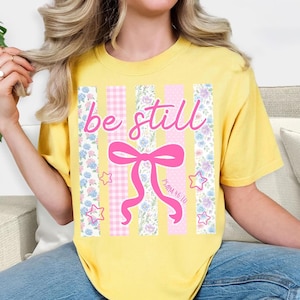 K&ouml;nnte beinhalten: Gelbes T-Shirt mit dem rosa Schriftzug "be still". Das Design beinhaltet eine rosa Schleife, Sterne und vertikale Streifen mit Blumen- und Vichy-Mustern. Der Text "Psalm 46:10" ist ebenfalls enthalten.