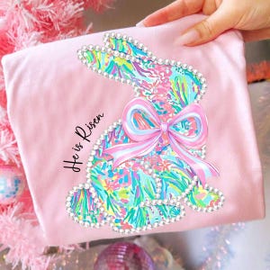 May include: Pink t-shirt featuring a colorful bunny design with a pearl border and a pink bow. The bunny is filled with a floral pattern in shades of blue, green, and pink. The text "He is Risen" is printed on the shirt.