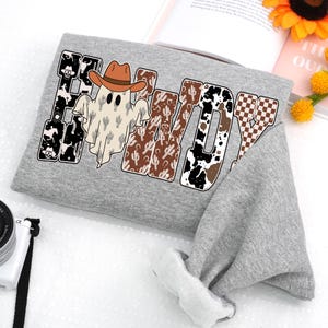 K&ouml;nnte beinhalten: Ein graues Sweatshirt mit dem Wort "Howdy" in einem Western-Design. Das Design umfasst einen Geist mit Cowboyhut, Kaktusmuster und Kuhfell-Akzente. Das Sweatshirt ist gefaltet, mit dem &Auml;rmel sichtbar.