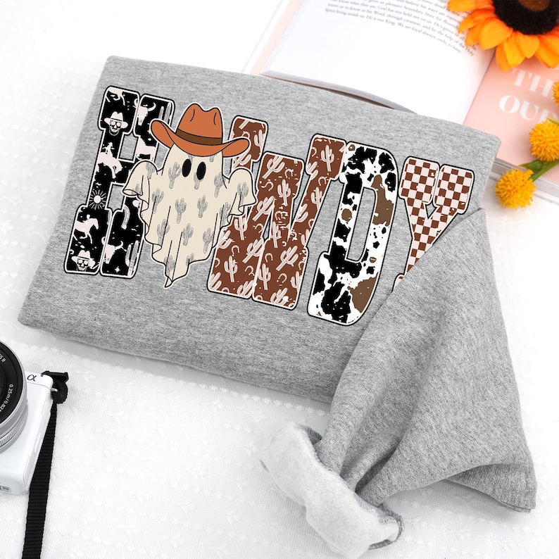 K&ouml;nnte beinhalten: Ein graues Sweatshirt mit dem Wort "Howdy" in einem Western-Design. Das Design umfasst einen Geist mit Cowboyhut, Kaktusmuster und Kuhfell-Akzente. Das Sweatshirt ist gefaltet, mit dem &Auml;rmel sichtbar.