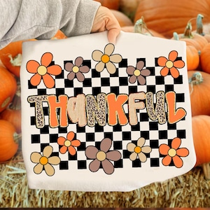 May include: White t-shirt with a colorful floral design and the word "THANKFUL" in orange and leopard print. The design includes orange, brown, and tan flowers, set against a black and white checkered background. A festive fall-themed graphic.