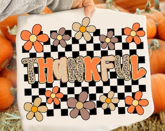 May include: White t-shirt with a colorful floral design and the word "THANKFUL" in orange and leopard print. The design includes orange, brown, and tan flowers, set against a black and white checkered background. A festive fall-themed graphic.