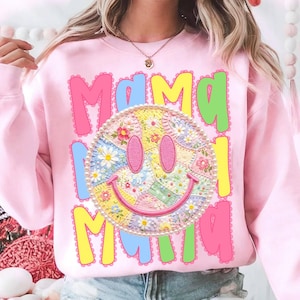 May include: Pink sweatshirt with the word "Mama" in colorful letters. A smiley face with a floral patchwork design is in the center, surrounded by a pearl border. The sweatshirt is a casual, comfortable style.