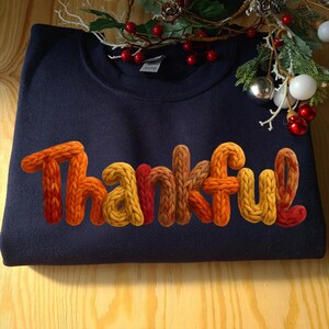 May include: A navy blue sweatshirt with the word "Thankful" in a 3D knit-style font. The letters transition from orange to yellow and red. Festive holiday decor is in the background.