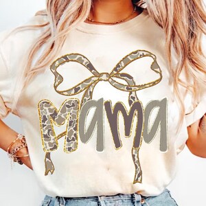 May include: Cream-colored t-shirt featuring the word "MAMA" in a gray, outlined font. A decorative bow with a camouflage pattern and gold trim is above the word. The shirt is a casual, everyday wear item.