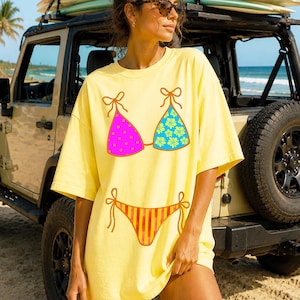 以下が含まれることがあります： ビキニのデザインが施された黄色のTシャツ。ビキニの上部は、紫のドットが入ったピンク色と、緑の花柄が入った青色です。下部はオレンジと黄色のストライプです。ビーチの近くで人が着ています。