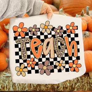 May include: Cream-colored tote bag with the word "TEACH" in orange and leopard print lettering. The bag features a black and white checkered pattern and is decorated with orange, brown, and cream-colored flowers. The background includes pumpkins.