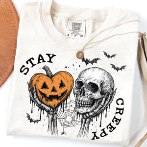 May include: Cream-colored t-shirt with a Halloween-themed graphic. The design features a jack-o'-lantern, a skull, bats, and the words "STAY CREEPY". A gold necklace with a pendant is also visible.