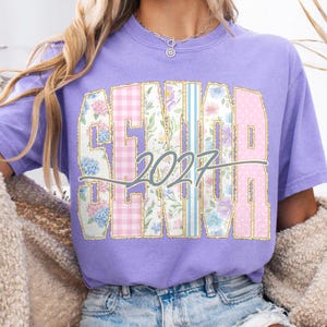 May include: A lilac-colored t-shirt featuring the word "SENIOR" in a floral and gingham pattern, with the year "2027" in a script font. The shirt has a classic crew neck and short sleeves, perfect for casual wear.