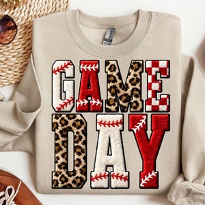 May include: Beige sweatshirt with the words "GAME DAY" in a baseball-themed design. The letters are a mix of baseball stitching, leopard print, and red and white checkered patterns. A casual, sporty top.