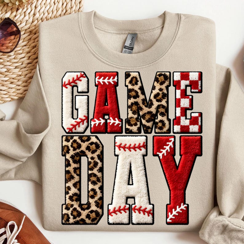May include: Beige sweatshirt with the words "GAME DAY" in a baseball-themed design. The letters are a mix of baseball stitching, leopard print, and red and white checkered patterns. A casual, sporty top.