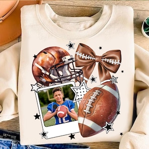 Könnte beinhalten: Cremefarbenes Sweatshirt mit Football-Motiv. Das Design umfasst einen braunen Footballhelm, einen Football, eine Schleife, ein Foto einer Person, die einen Football hält, und Sternakzente.