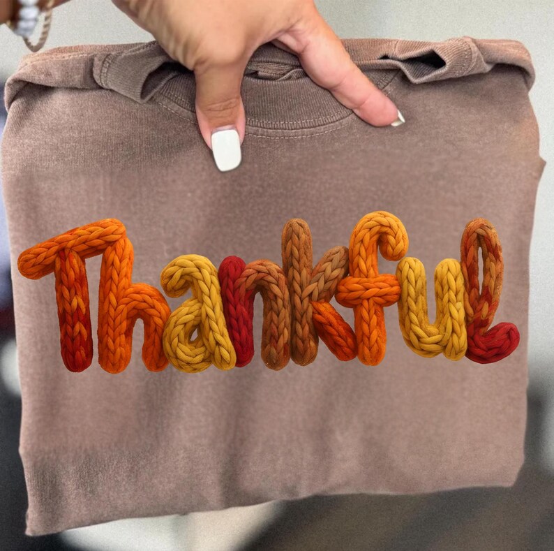 May include: A brown long-sleeve shirt with the word "Thankful" in colorful, textured, knitted letters. The letters transition from orange to yellow and red. The shirt is held by a hand.