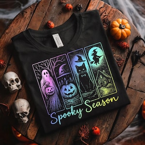 May include: Black t-shirt with a colorful "Spooky Season" graphic. The design features a ghost, a pumpkin, a black cat, and a witch flying in front of a full moon. The shirt is folded on a wooden surface with Halloween decorations.