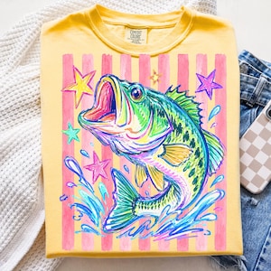 Op de afbeelding: Geel T-shirt met een kleurrijke afbeelding van een springende baars, omringd door sterren en waterspetters. Het ontwerp heeft roze verticale strepen. Het T-shirt is op een wit oppervlak gevouwen.
