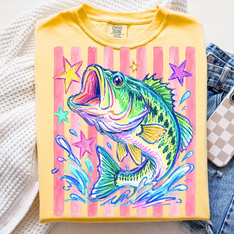 Op de afbeelding: Geel T-shirt met een kleurrijke afbeelding van een springende baars, omringd door sterren en waterspetters. Het ontwerp heeft roze verticale strepen. Het T-shirt is op een wit oppervlak gevouwen.