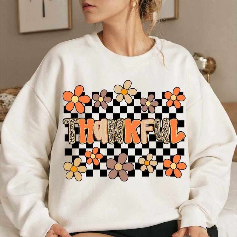 May include: Cream-colored sweatshirt with the word "THANKFUL" in orange and leopard print lettering. The design includes a black and white checkered background and colorful flower graphics. A casual and festive fall-themed top.
