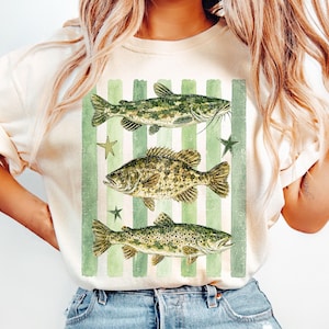 May include: Cream-colored t-shirt featuring a vintage-style illustration of three fish: a catfish, a bass, and a trout. The design includes green vertical stripes and star accents, creating a rustic, outdoorsy aesthetic. The fish are detailed with realistic textures and colors.