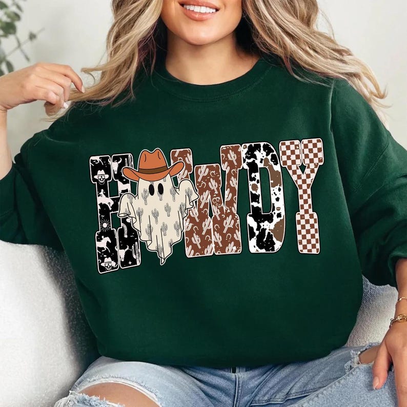 K&ouml;nnte beinhalten: Dunkelgr&uuml;nes Sweatshirt mit dem Wort "Howdy" in einem Design im Western-Stil. Der Buchstabe "H" zeigt einen Geist mit Cowboyhut. Die Buchstaben haben Muster wie Kuhfell, Kakteen und ein braun-wei&szlig;es Schachbrettmuster.