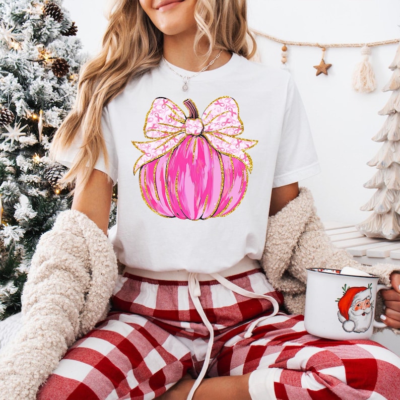 May include: White t-shirt featuring a pink pumpkin design with a gold glitter outline and a matching bow. The bow is adorned with pink ribbons. The person is wearing red and white plaid pajama pants and holding a mug with a Santa Claus design.