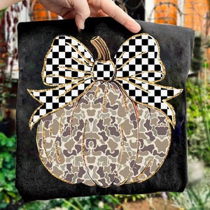 May include: Black square pillow featuring a pumpkin design. The pumpkin has a camouflage pattern in shades of brown and gray, with a gold glitter outline. A large bow with a black and white checkered pattern sits atop the pumpkin.