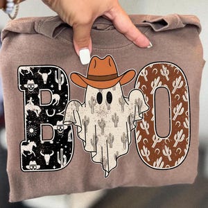 May include: A brown t-shirt featuring a Halloween design. The word "BOO" is displayed with a cowboy ghost in the middle. The letters are decorated with cacti, skulls, and other western-themed images.